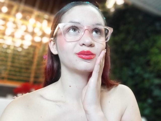 Lesley Gray, avec ses lunettes roses, son rouge à lèvres rouge et sa main sur sa joue, a les cheveux teints en rouge, une peau pâle et de petites taches de rousseur sur son visage, et elle regarde vers le haut avec une expression mignonne..