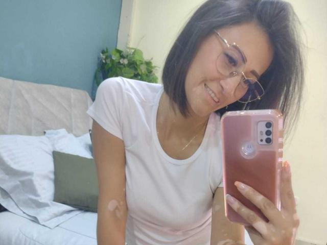SharissWilliam with short black hair, glasses, white tee, small bandage on arm, smiling at phone, pink case, holding it with right hand, reflection in mirror.