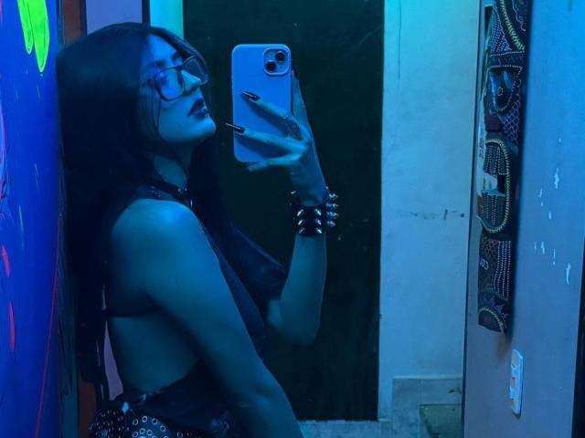 DarkMaddy with long black hair, glasses, and dark lipstick, taking a mirror selfie with blue lighting. She's wearing a black lace top, showing her bare side-boob.