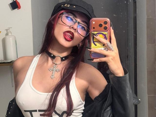 DarkMaddy with red lipstick, tongue out, wearing glasses, black cap, white tank top, choker, cross necklace, and black jacket, holding phone, showing off her tits and ass.