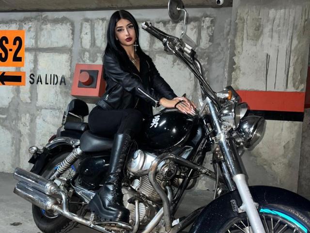 DarkMaddy with long black hair, wearing a black leather jacket and boots, sitting on a shiny black motorcycle, hands on the handlebars, in a gritty, concrete-walled garage.