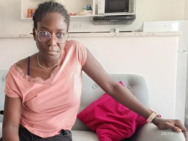 NerySkiraa has dark skin, small tits, wearing a pink shirt with lace, glasses, and a bracelet, sitting on a gray sofa with a pink pillow, looking at the camera.