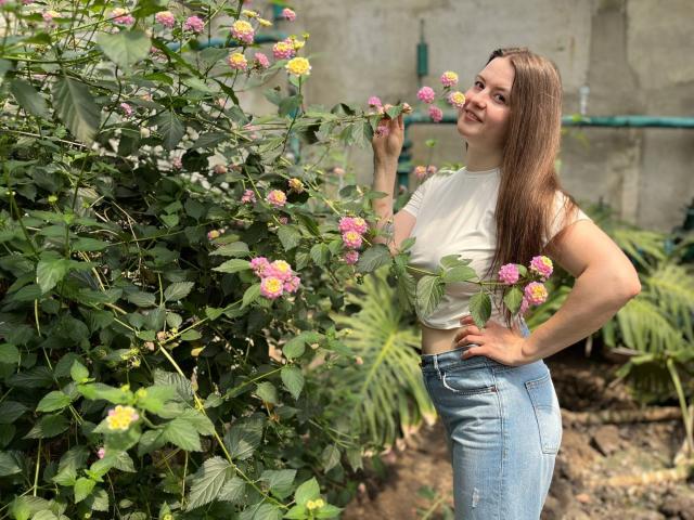 Mavka, con cabello castaño largo, una camiseta blanca y jeans de cintura alta, sonriendo a flores rosas y amarillas, sosteniendo una en su mano, de pie junto a follaje verde..