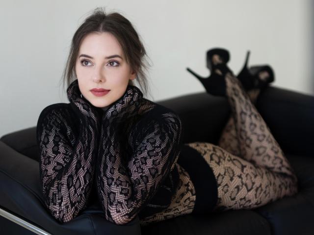 SophieXLips lies on a black leather couch, wearing a black lace top and leopard print leggings, with black high heels, her tits partially covered, legs up, looking at camera.