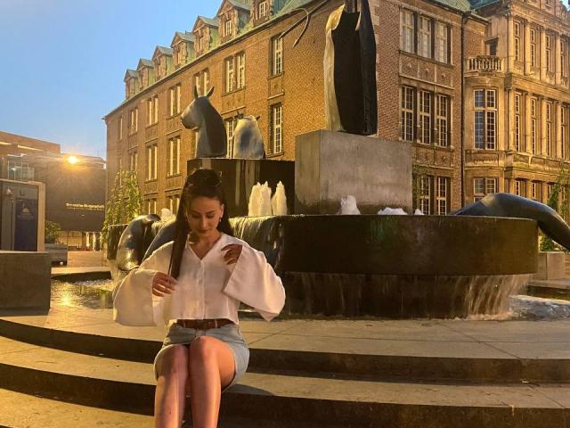 DominikaMorgan sits on stone steps, wearing a white blouse and blue skirt, adjusting her brown belt. She has dark hair in a bun, looking down. Water fountain behind.