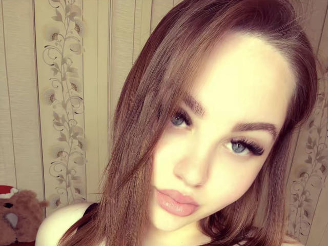 LydaMearns has light brown hair, blue eyes, and pink lips. She's looking at the camera with a slight pout, wearing black eyeliner and a white top.