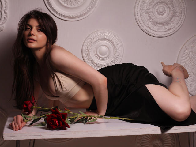 StellaLewin is lying on a white table, wearing a beige bra and black skirt, with red roses, showing her ass and bare legs, looking seductively at the camera.