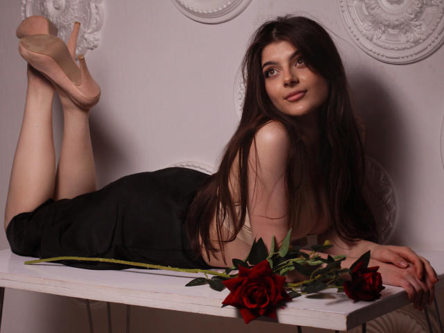 StellaLewin, lying on her stomach, black skirt, nude heels, long dark brown hair, red roses on white table, legs up, smiling, pale skin, small tits, cute face, relaxed.