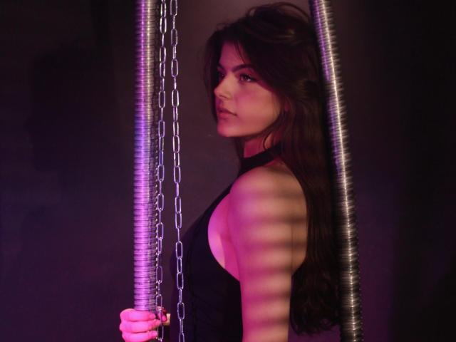 StellaLewin with long brown hair, side profile, wearing a black sleeveless top, holding metal chains, purple and pink lighting, looking serious, striped shadows on her arm.