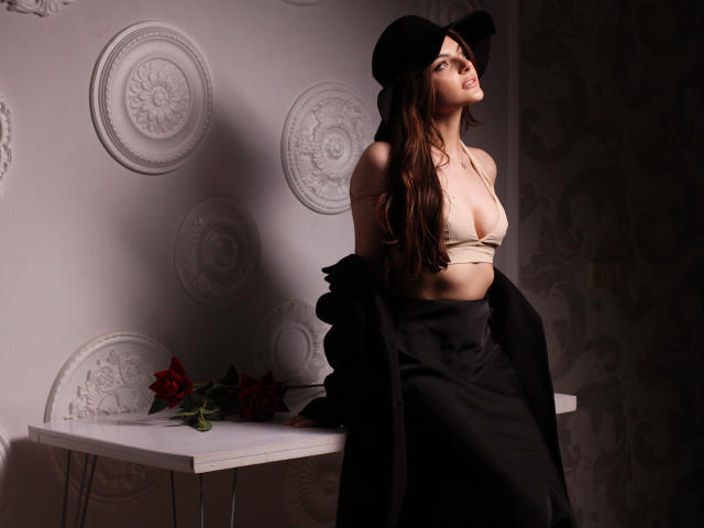 StellaLewin with long brown hair, wearing a black hat, beige bra showing her tits, black coat off, standing by a table with red roses, looking up.