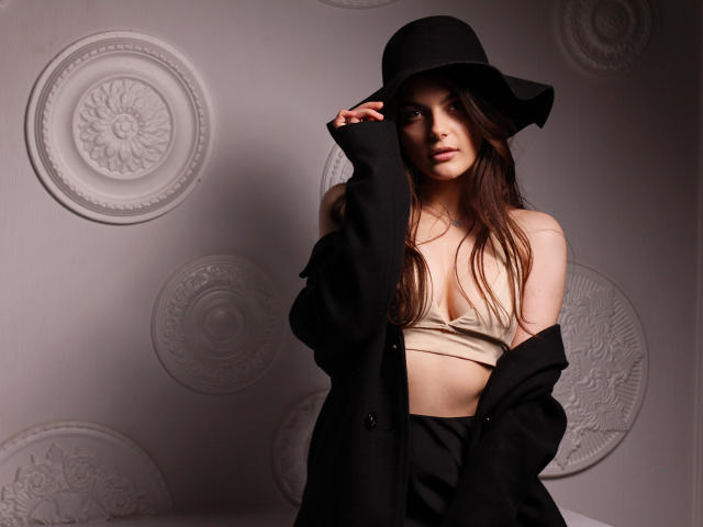 StellaLewin with long brown hair, wearing a black hat, off-shoulder black coat, and beige bra, exposing her tits, standing against a patterned wall, looking seductively at camera.