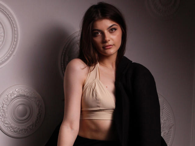 StellaLewin with medium tits in a beige sports bra, dark brown hair, black jacket, and a serious expression, standing against a patterned wall.