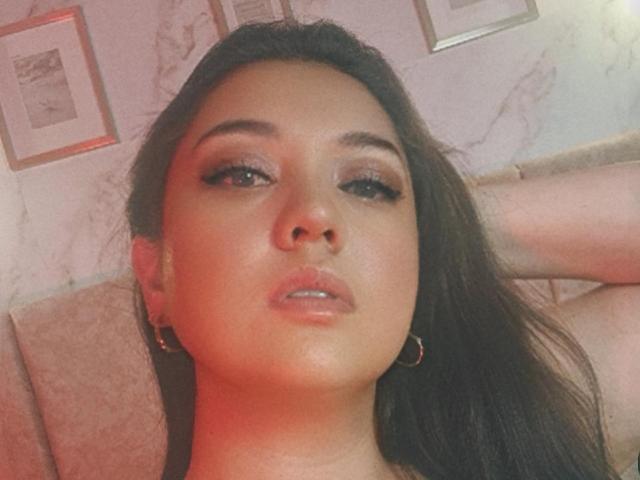 PaulaAnd has dark brown hair, slightly parted lips, and wears small gold hoop earrings. Her makeup includes smoky eyeshadow and winged eyeliner. Her skin is light brown.