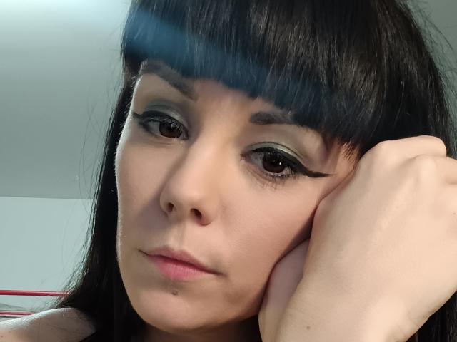 OrianaLaFrancaise with dark straight hair, blue-green eyeshadow, black winged eyeliner, and pink lips, resting her chin on her hand, looking serious. Her skin is light and smooth.