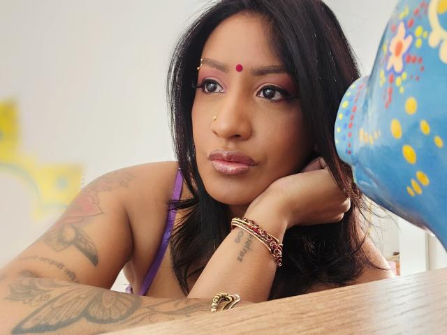 Nahirr with dark hair, brown skin, red bindi, pink eye makeup, tattoos on arm, purple top, gold nose ring, gold bracelet, resting chin on hand, wooden table.