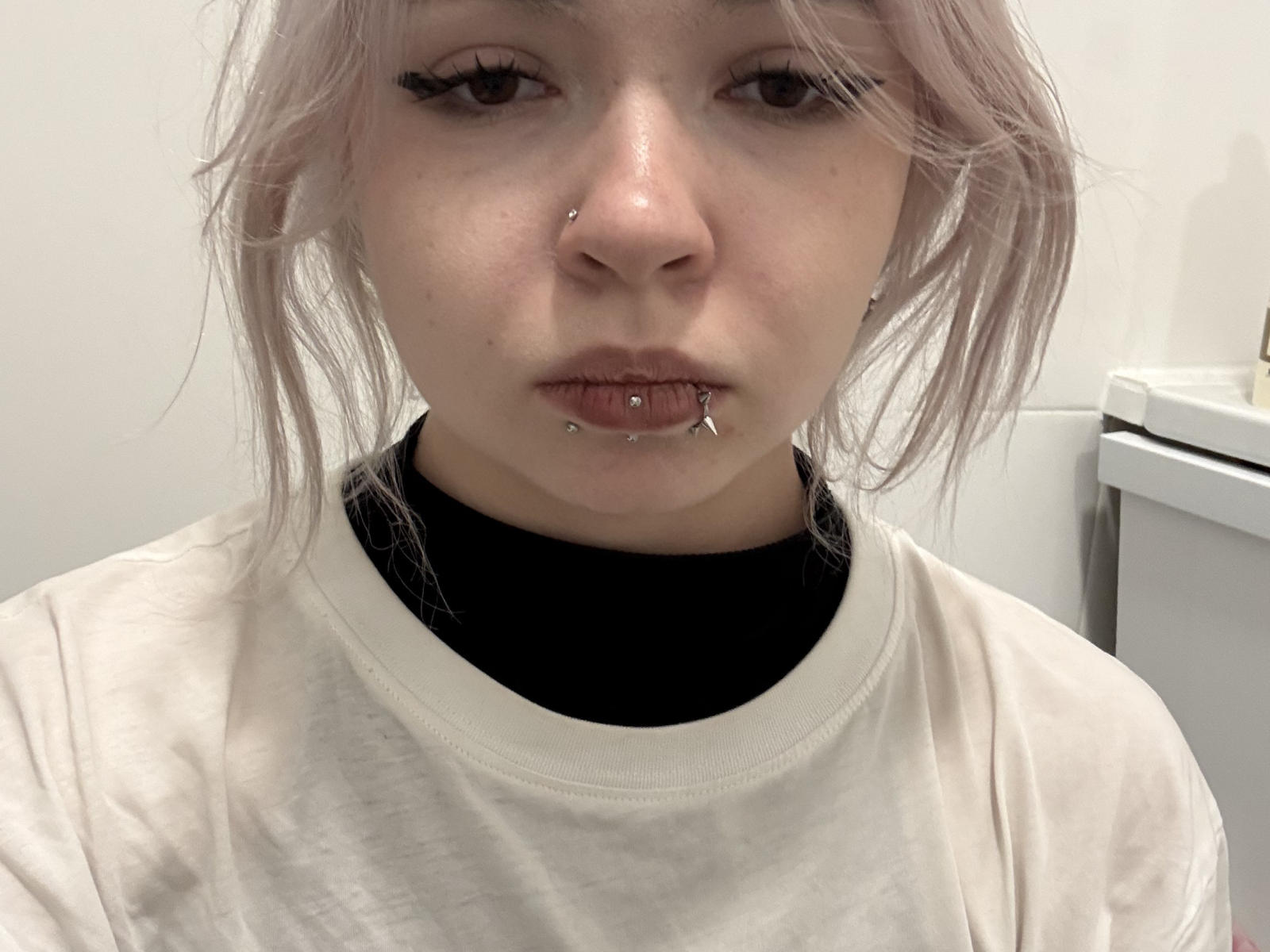 MaryannaGoines has pale pink hair, a nose ring, and a lip piercing with a small cross. She's wearing a black turtleneck and a white shirt.