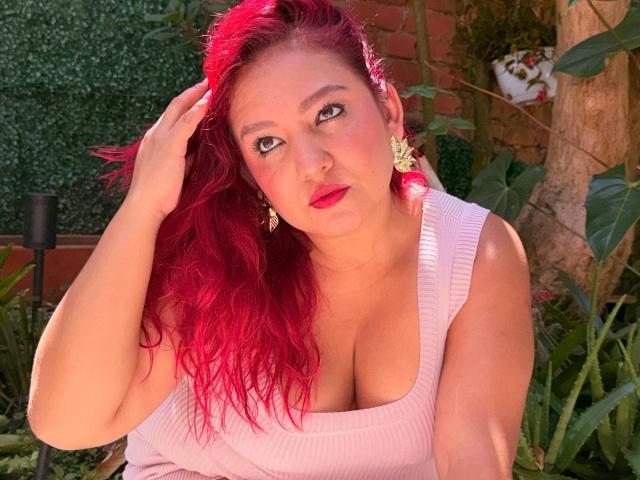 DorothyMoon with bright red hair, wearing a pink ribbed top, showing her big boobs, pink lipstick, gold earrings, hand in hair, green-eyed, sitting among plants.