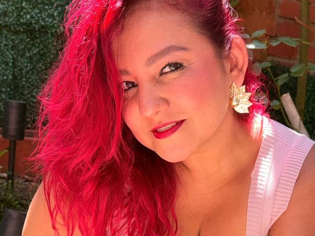 DorothyMoon with bright red hair, white ribbed tank top, and star earrings, smiling with red lipstick, showing her tits, nose ring, and freckles, in a sunny garden.