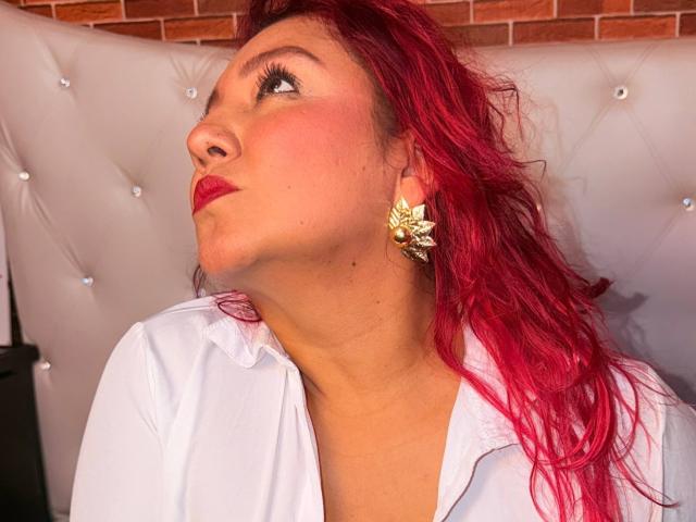 DorothyMoon with red hair, pouty lips, and gold leaf earrings, looking up with her ass slightly raised, wearing a white shirt, in a tufted, white headrest.