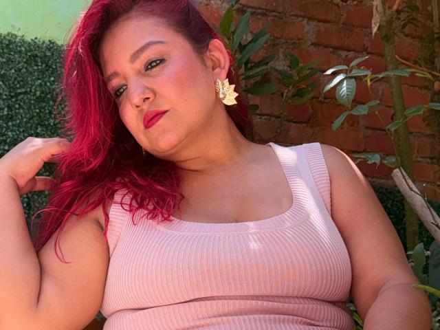 DorothyMoon with bright red hair, pink ribbed tank top, and yellow flower earrings, showing off her tits, hand in hair, red lipstick, and a confident, slightly smug expression.