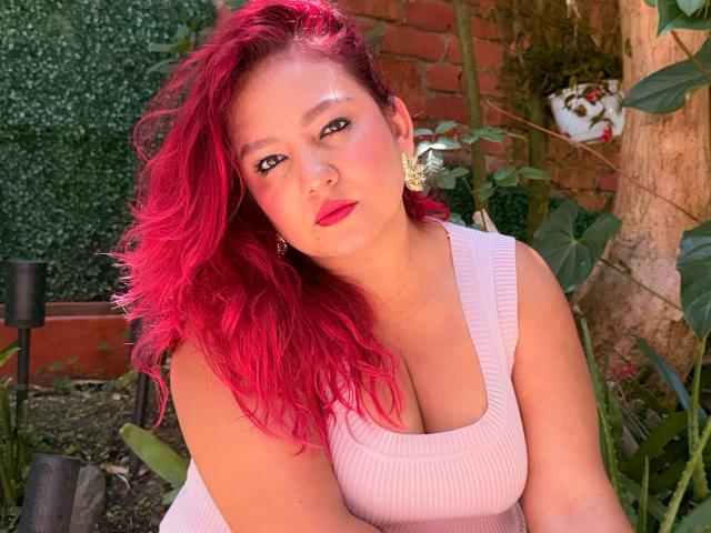 DorothyMoon with bright red hair, pink tank top, and gold earrings, sitting outdoors, showing her tits, with a serious expression, and red lipstick.