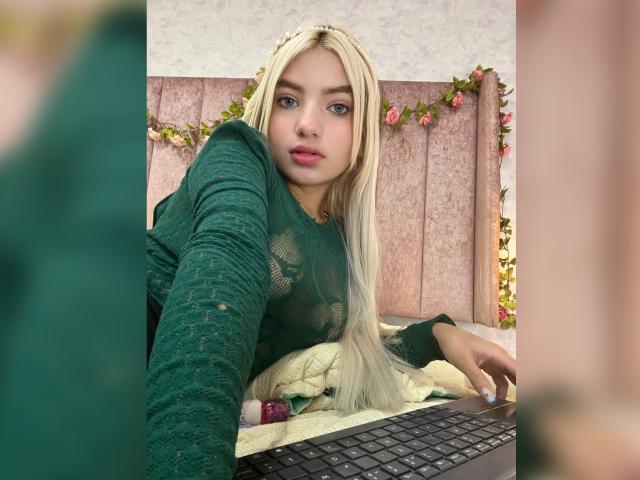 SabrinaColle mit blonden Haaren, einem grünen Spitzenoberteil und pinkem Lippenstift, macht ein Selfie, während sie auf einem Laptop tippt. Ihr süßes Gesicht und ihre kleinen Brüste sind auf dem Foto zu sehen..