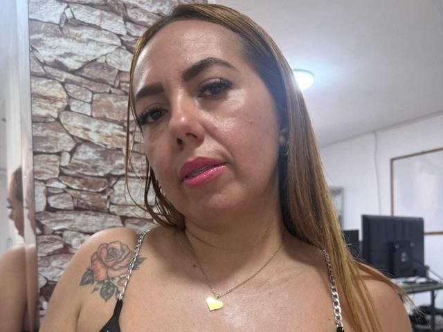 AmberDillucca with tan skin, pink lipstick, and brown hair, wearing a black top with a rose tattoo on her right shoulder, a heart necklace, and looking at the camera.