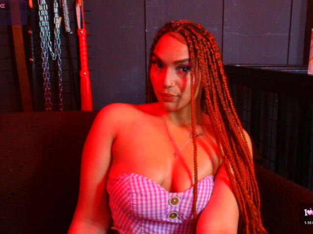 BrianaPratt with long braided red hair, wearing a pink checkered top that shows her big tits, sitting in a red-lit room, looking at the camera.