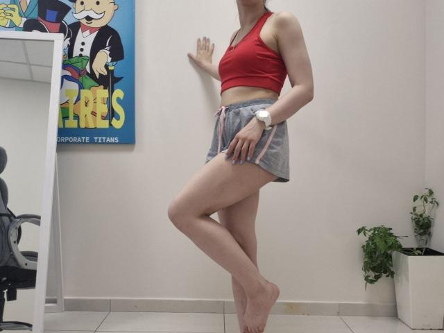 JoannaSia se tient pieds nus, vêtue d'un soutien-gorge de sport rouge et d'un short gris, sa main droite appuyée contre le mur, la jambe gauche croisée, exposant ses fesses et une montre à son poignet..
