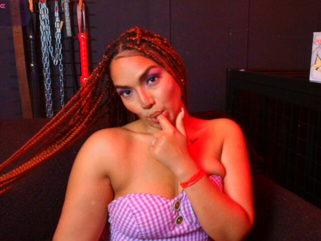 BrianaPratt with long braids, purple eye makeup, and a pink checkered top, touching her finger to her lips, showing her tits, in a red-lit room.