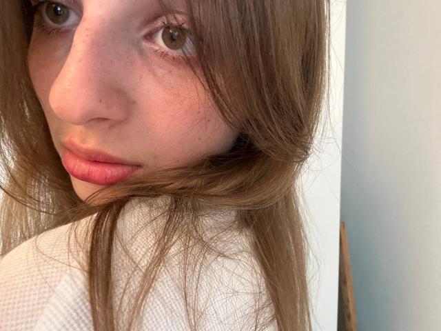 Le visage de KarieMestler, avec ses yeux marron et ses lèvres roses, est en premier plan. Elle porte un haut blanc texturé, et ses longs cheveux bruns recouvrent un côté de son visage. On peut voir ses taches de rousseur..