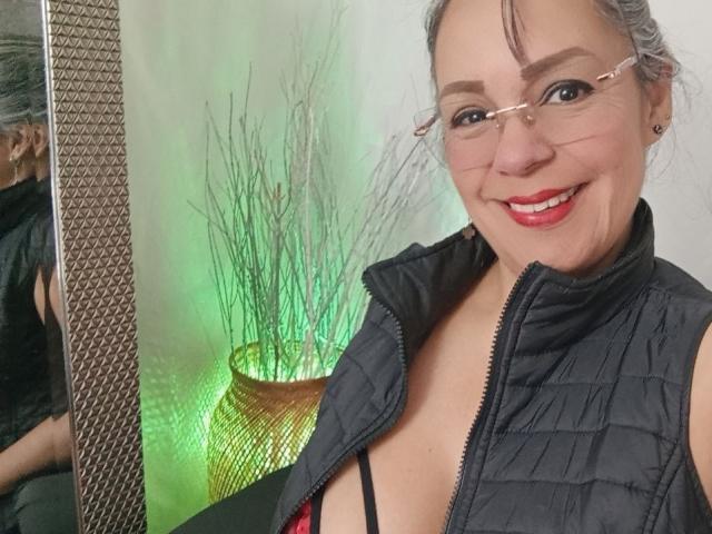 Laila Castillo, avec des lunettes, souriante, portant un gilet noir matelassé, avec une partie de sa poitrine visible, du rouge à lèvres rouge et un soutien-gorge noir. Son reflet dans le miroir montre son dos. Une lumière verte est visible en arrière-plan..