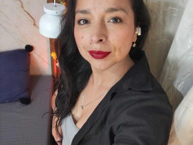 StangelarRivera with dark hair, red lipstick, black shirt, gray tank top, small tits, white earrings, necklace, smiling, brown eyes, fair skin, in a cozy indoor setting.
