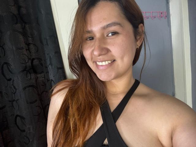 SofiaVelvet ha lunghi capelli castani, sta sorridendo e indossa un top nero senza spalline che mette in mostra le spalle e la parte superiore del petto, con evidenti segni della abbronzatura e piccoli orecchini..
