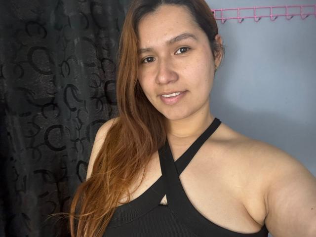 SofiaVelvet, con lunghi capelli castani, indossa un top nero a fascia che mette in mostra il seno, ha la pelle chiara, piccoli orecchini, un leggero sorriso e sta in piedi di fronte a una tenda scura..