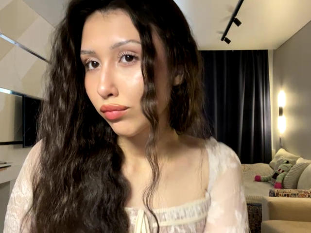 Necole Arciniega, met golvend zwart haar, lichte huid en roze lippen, draagt een witte kanten top en kijkt neutraal naar de camera. De borsten zijn licht zichtbaar..