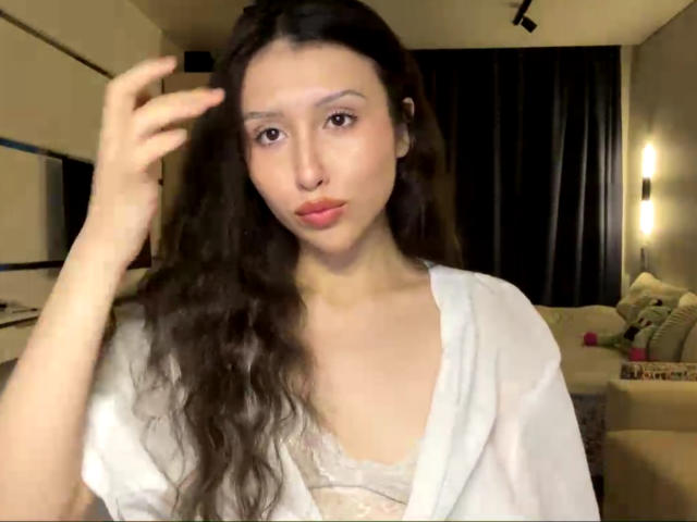 NecoleArciniega, con cabello castaño largo y ondulado, una camisa blanca abierta y labios rosados, tocándose el cabello, mirando a la cámara, mostrando senos pequeños, en una habitación con poca luz..