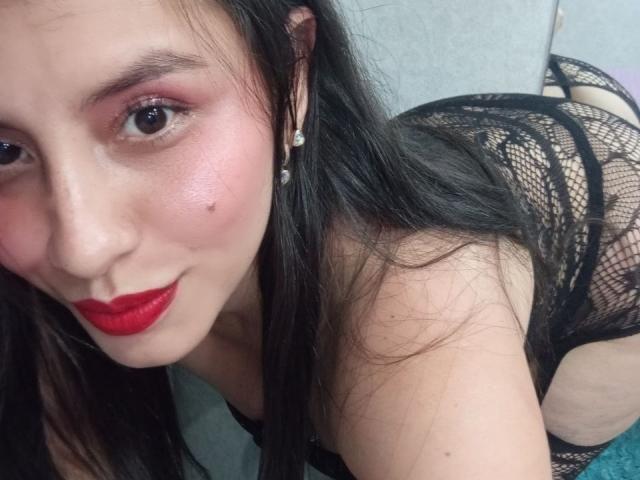 MadissonFontaine, com lábios vermelhos, lingerie de renda preta e uma pequena pinta na bochecha, olhando para a câmera, exibindo suas nádegas e meias de rede..
