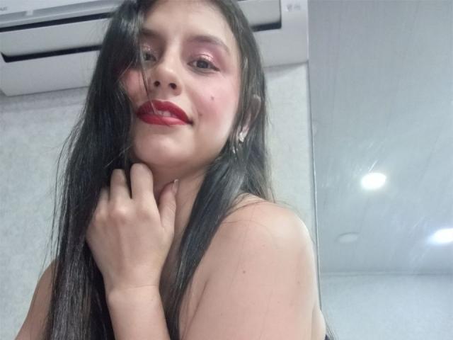 MadissonFontaine, com cabelos longos e pretos, batom vermelho, sombra rosa, sorrindo, mão perto do queixo, unidade de ar condicionado acima, parede branca, pequenas imperfeições na pele, pele clara, sem roupas visíveis..