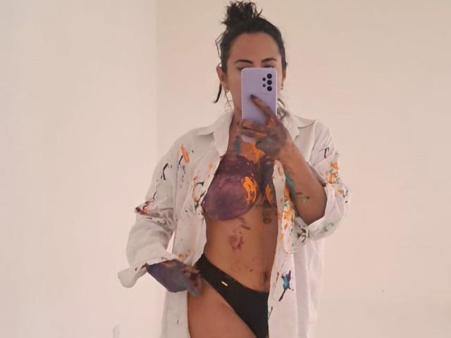 Jade Foster, mit Tattoos, trägt ein weißes, geblümtes Hemd, das offen ist, um ihre Brüste in einem lila BH und schwarzen Unterhosen zu zeigen, und macht ein Selfie vor einem Spiegel..