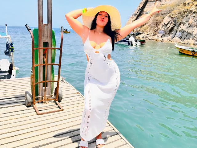 MiaDenver, formosa con un seno prosperoso e un sedere sexy, indossa un abito bianco con aperture e un cappello da sole, ed è in piedi su un molo di legno con le braccia alzate, vicino a delle barche..