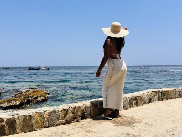 MiaDenver con la pelle scura, capelli neri, cappello bianco a tesa larga, top senza maniche, gonna bianca a vita alta, tacchi neri, in piedi su un sentiero di pietra, di fronte all'oceano, con barche nell'acqua..