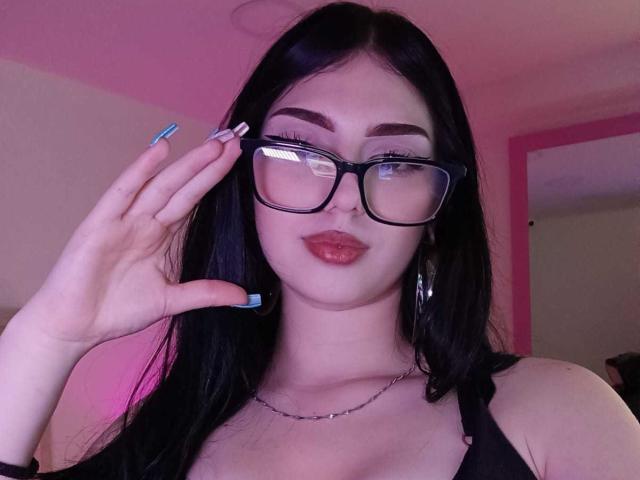 Isabella Rossi, com óculos pretos, unhas azuis e um colar, ajustando seus óculos, tem lábios vermelhos carnudos e cabelos longos pretos, mostrando seus seios em uma blusa preta..