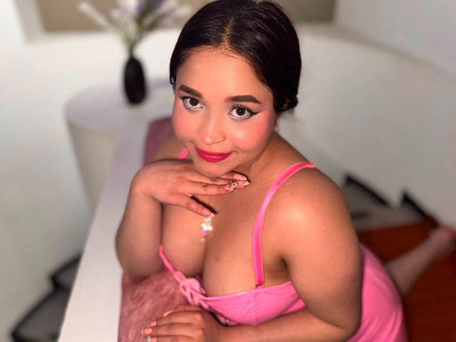 SamyMancini with big tits, wearing pink lingerie, has dark hair in a bun, pink lipstick, and is leaning on a white table, smiling with a hand near her mouth.