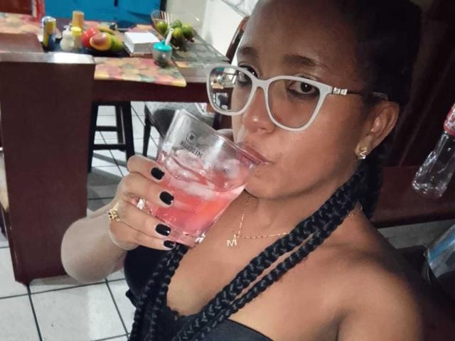 Lilith en Asmodeo, met donkere huid, gevlochten haar, witte bril, zwarte nagels, die een roze cocktail drinken, een gouden ketting dragen, met kleine borsten, in een slecht verlichte kamer, waarbij ze een glas in hun rechterhand houden..