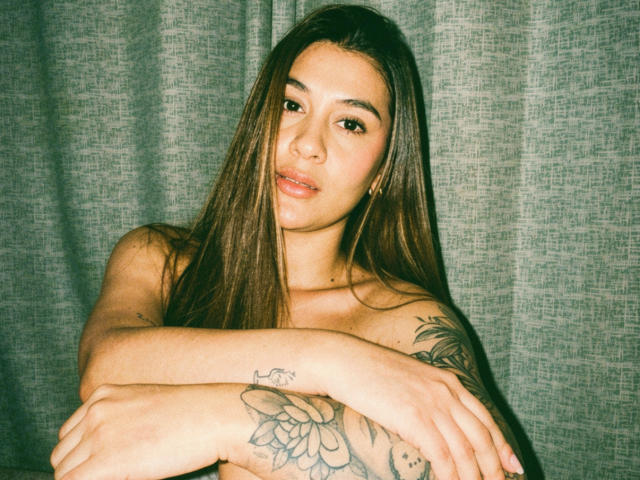 ThaliaAres, avec de longs cheveux bruns, les bras croisés, montrant une fleur tatouée sur l'avant-bras droit, une peau claire, un maquillage discret et un léger sourire, debout devant un rideau gris..