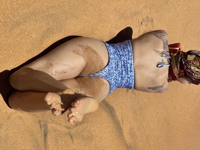 SarahRousen lying on sandy beach, wearing blue floral-patterned thong, butt and thighs covered in sand, tattoo on right side, feet in foreground, head adorned with colorful bandana.