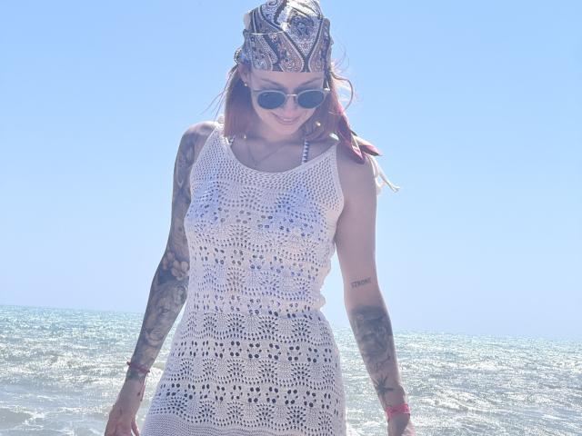 SarahRousen with tattoos, wearing a white, see-through, lace dress, bandana, and sunglasses, standing in the ocean, smiling, showing her tits through the dress.