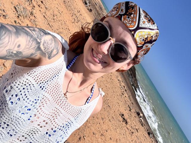 SarahRousen with tattoos, wearing a white mesh top, black-and-white patterned bandana, and dark sunglasses, smiling at the beach. Her tits are small, and she has red hair.