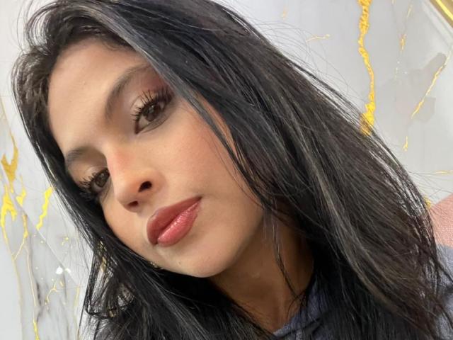NyraVanel a de longs cheveux noirs, des yeux marron avec un trait d'eye-liner prononcé, et des lèvres rouges brillantes. Sa peau est bronzée, et elle regarde légèrement sur le côté avec un sourire discret..